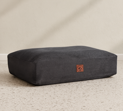 Dark gray rectangular pet bed with a brown logo on a light gray floor.