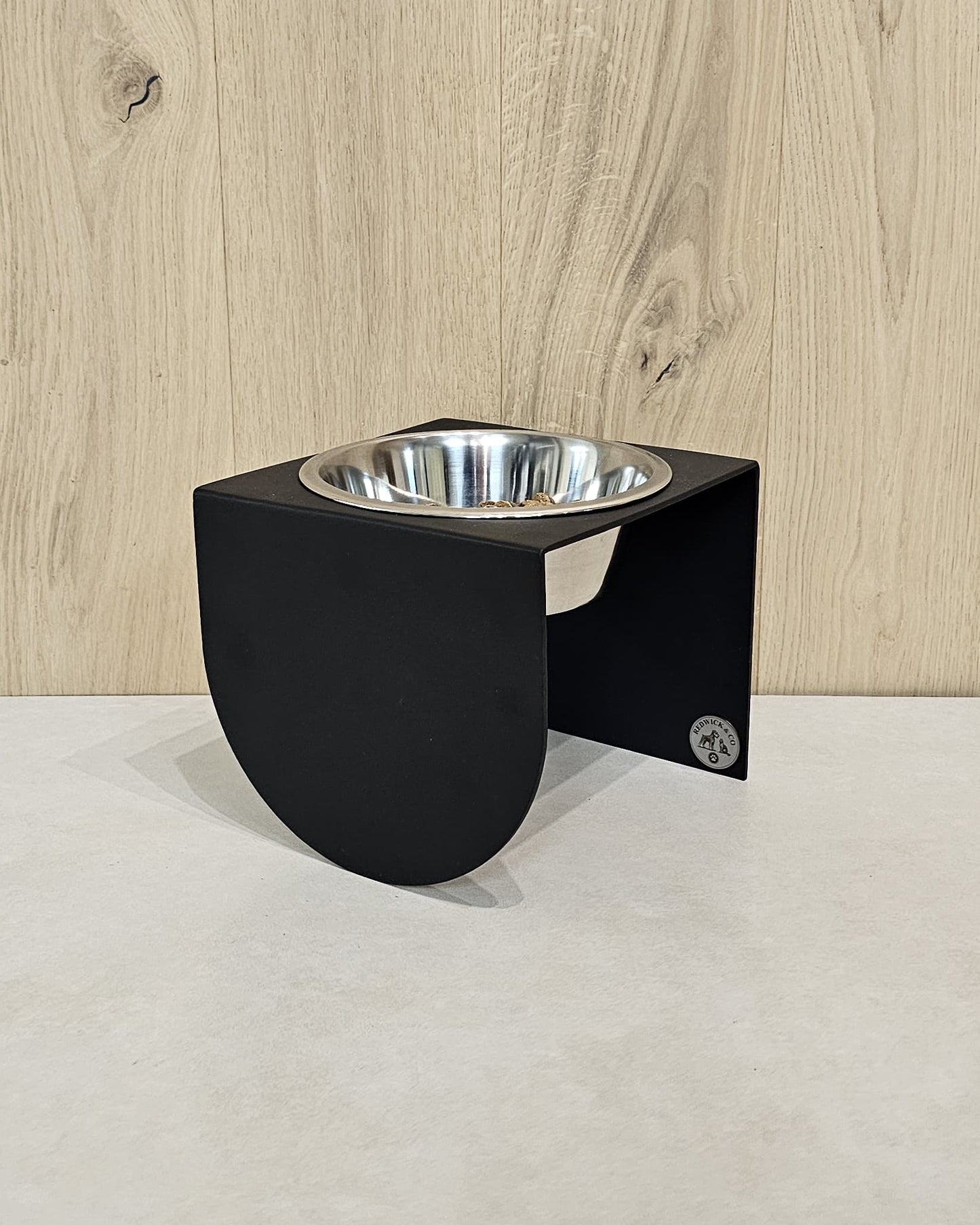Black elevated pet feeder with a silver bowl on a light wood floor.