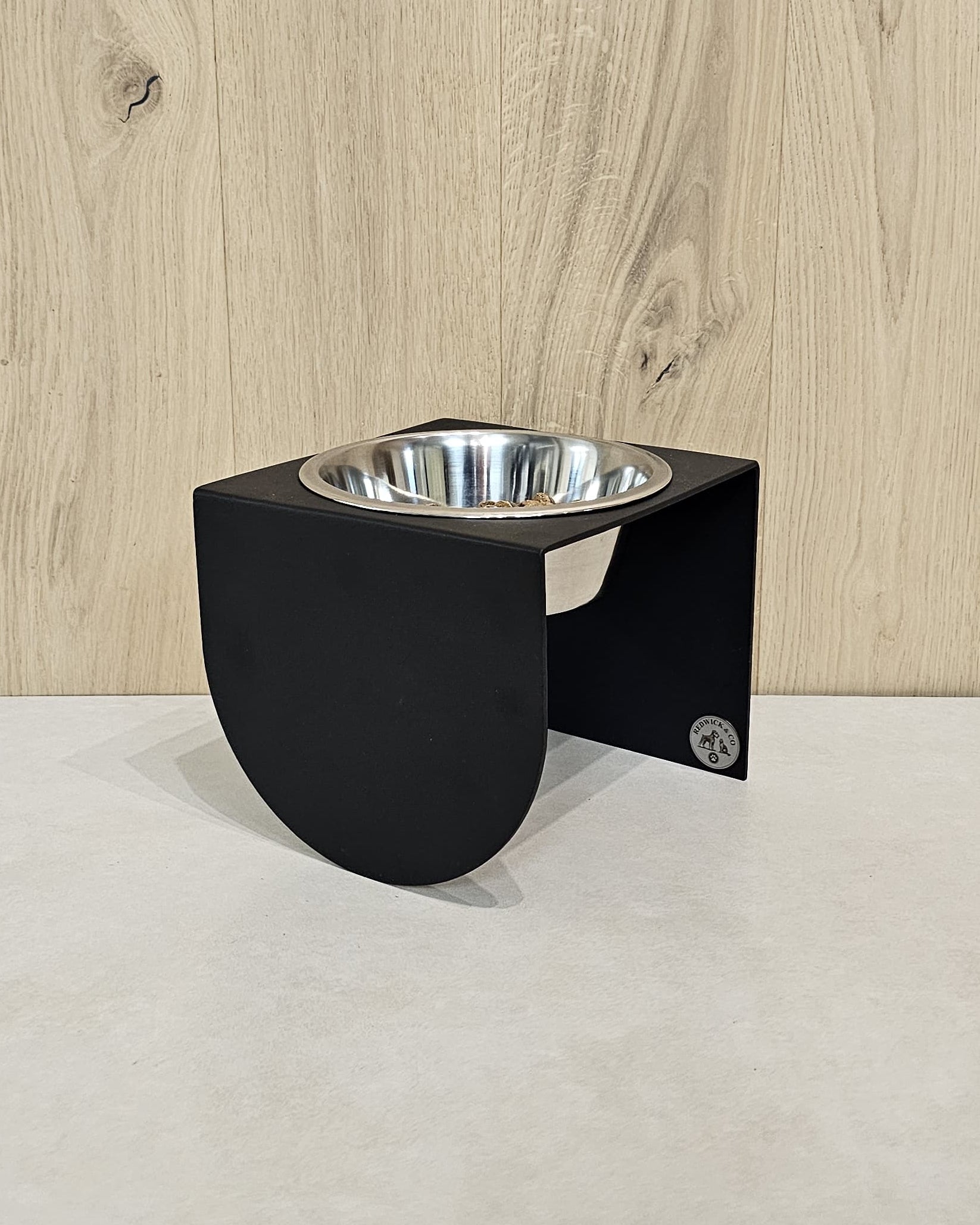 Black elevated pet feeder with a silver bowl on a light wood floor.